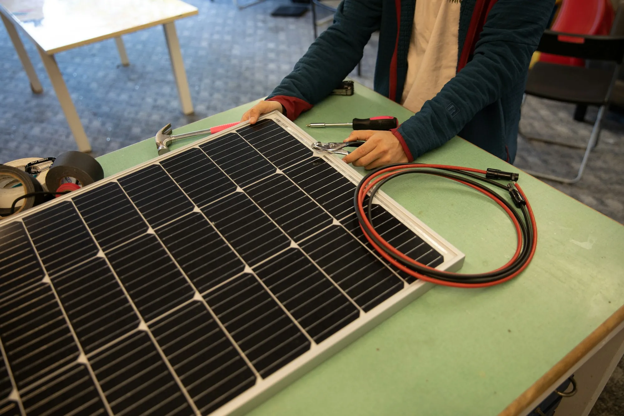 Hands working on a small solar panel and its red and black cable on a workbench