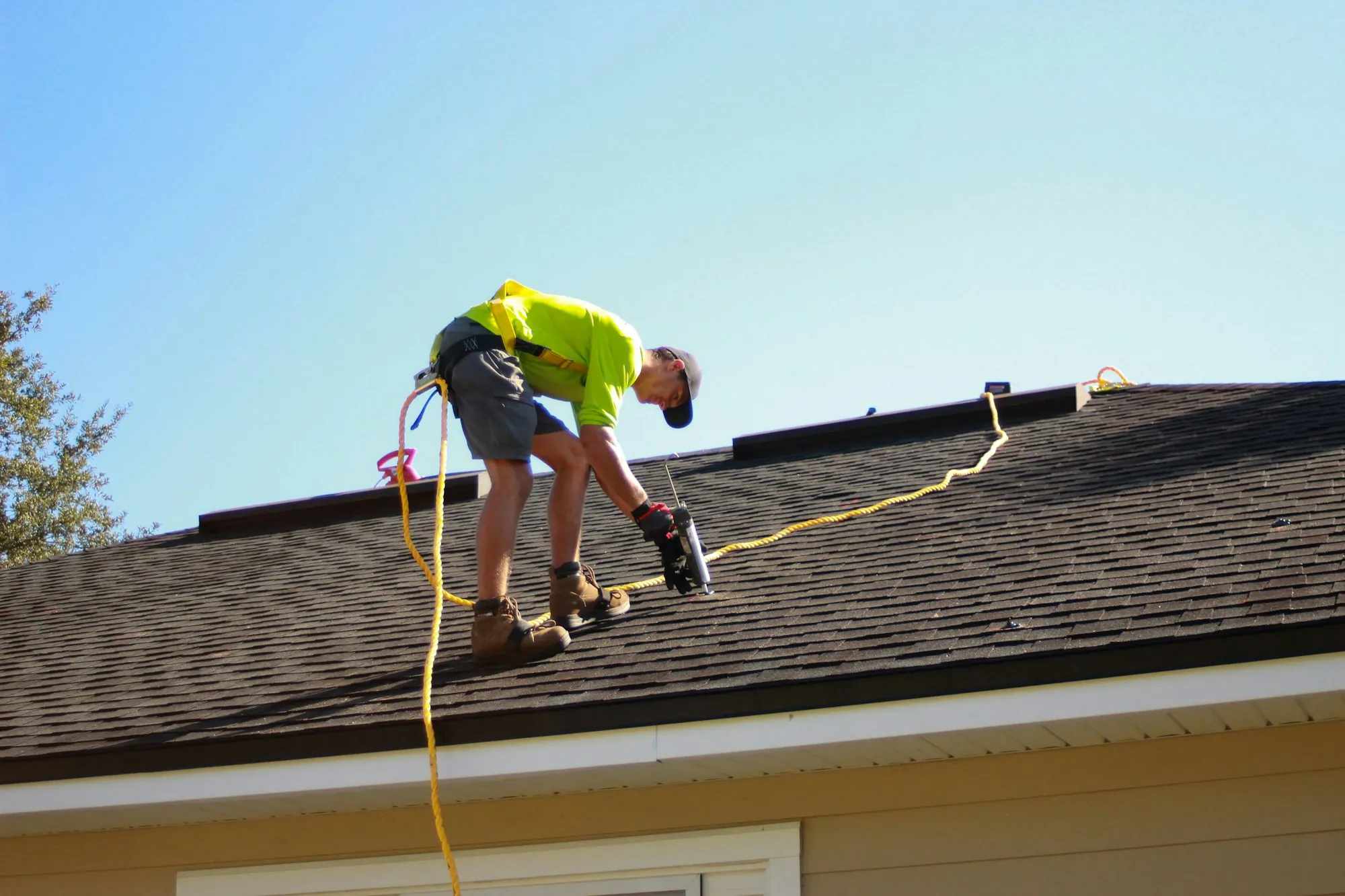 Roofer in a harness bent over working on a shingle roof against a blue sky